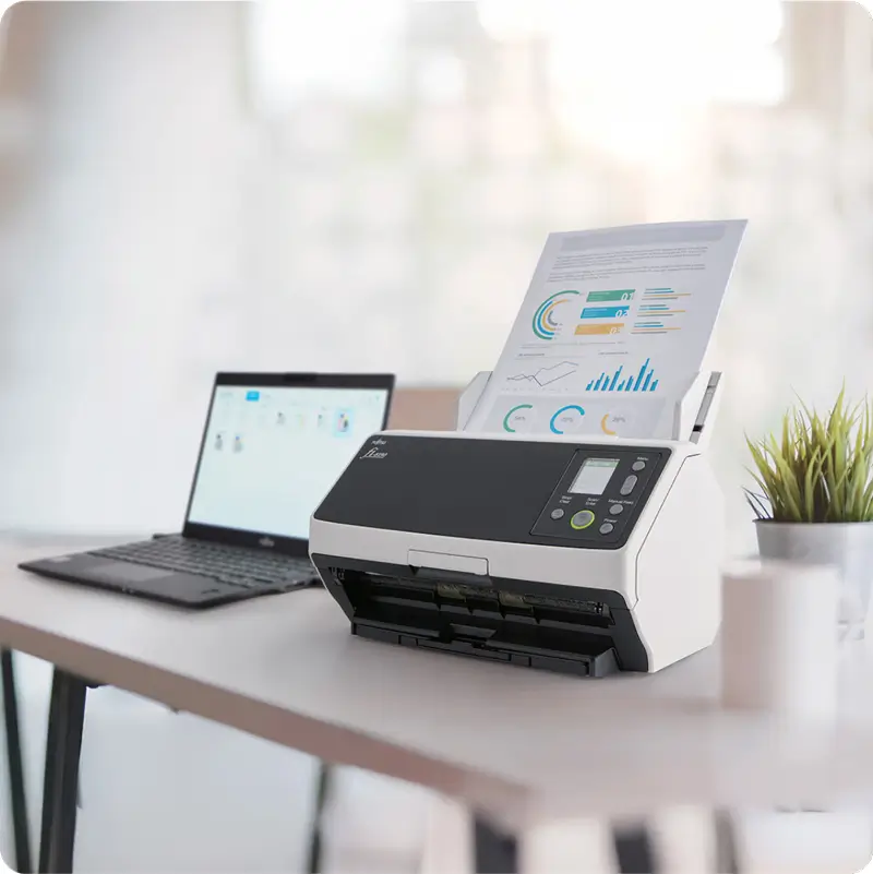 Document scanner on a desk in a minimalist office, processing a colorful report with graphs, next to an open laptop and a small plant."