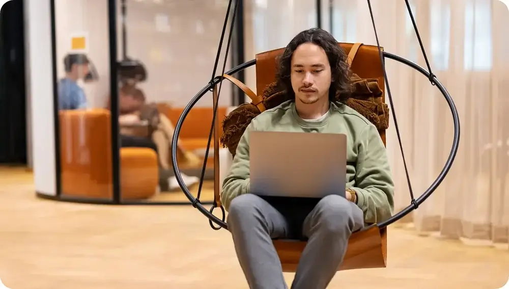 Man using a laptop while sitting in a hanging chair in a casual office setting.