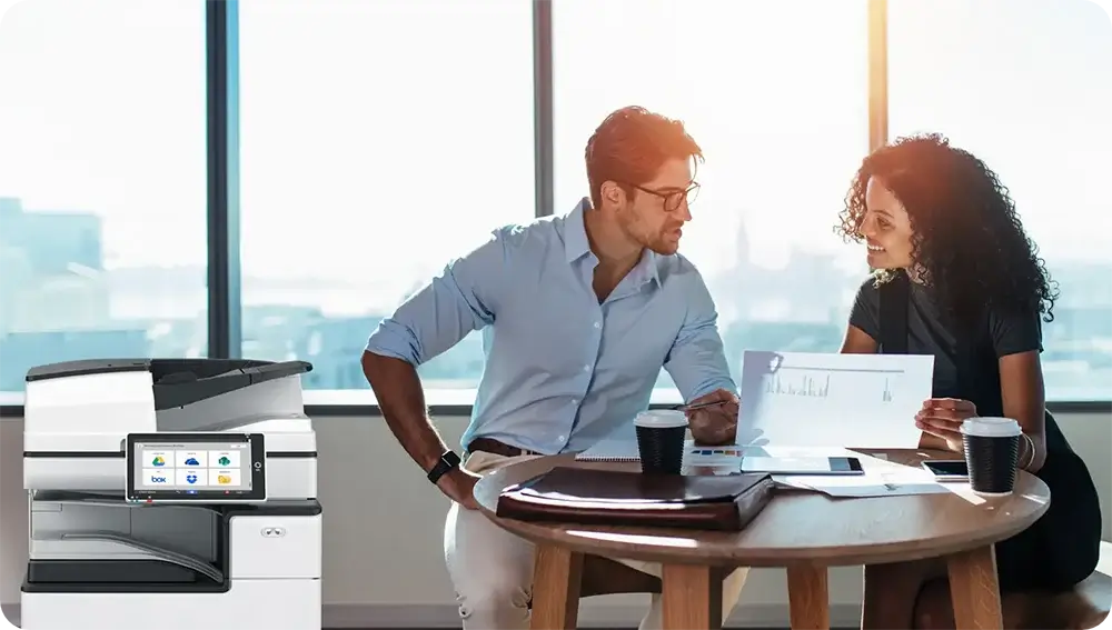 Two professionals discussing documents at a table in a bright office with city views.